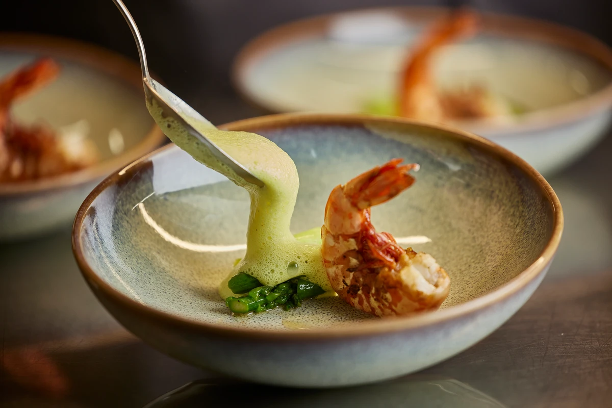 Nahaufnahme einer kunstvoll angerichteten, gebratenen Garnele mit etwas Spargel, die mit schaumiger Soße bedeckt werden